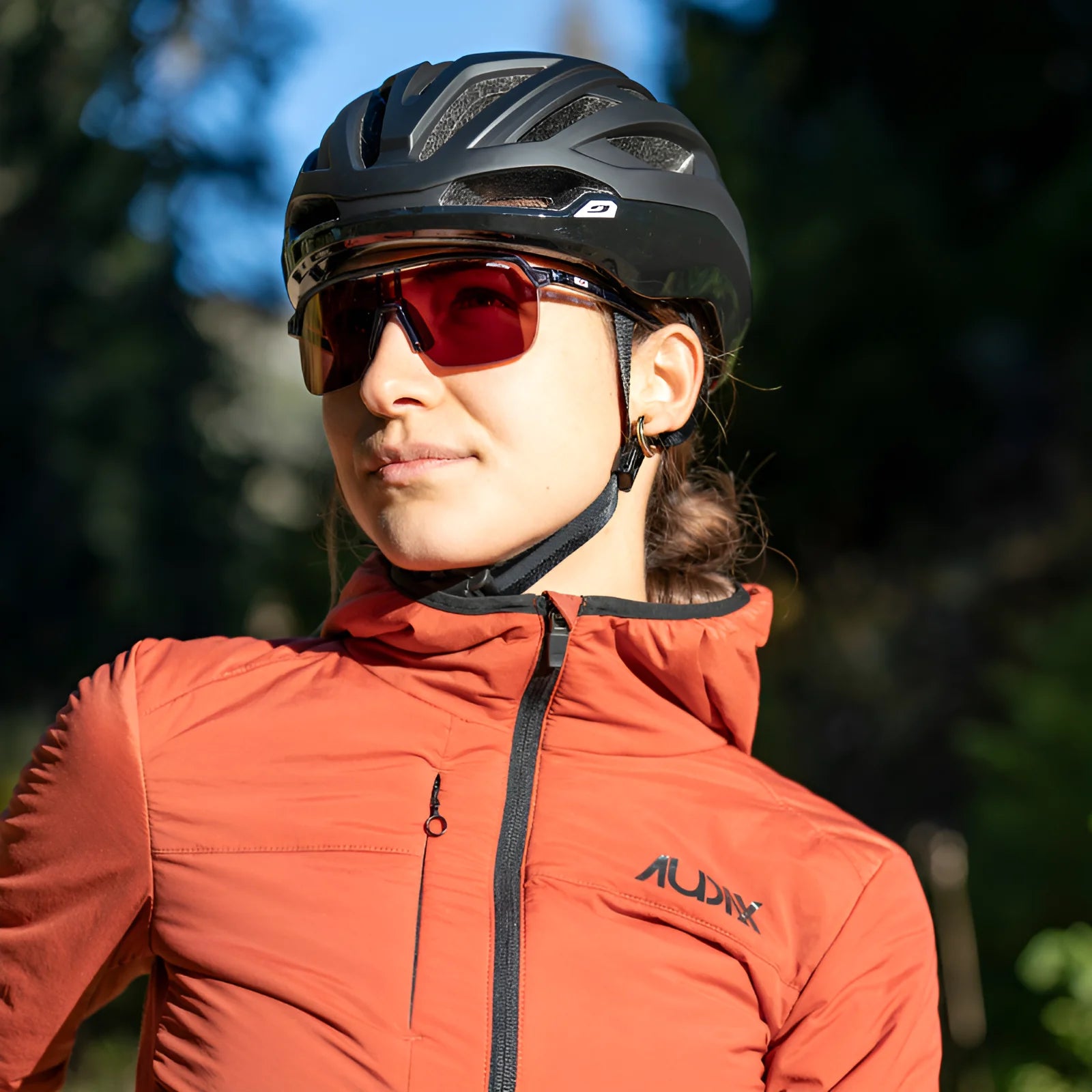 Wearing Julbo Frequency Sunglasses and a helmet, a person in an orange jacket stands ready for outdoor adventures against a blurred backdrop.