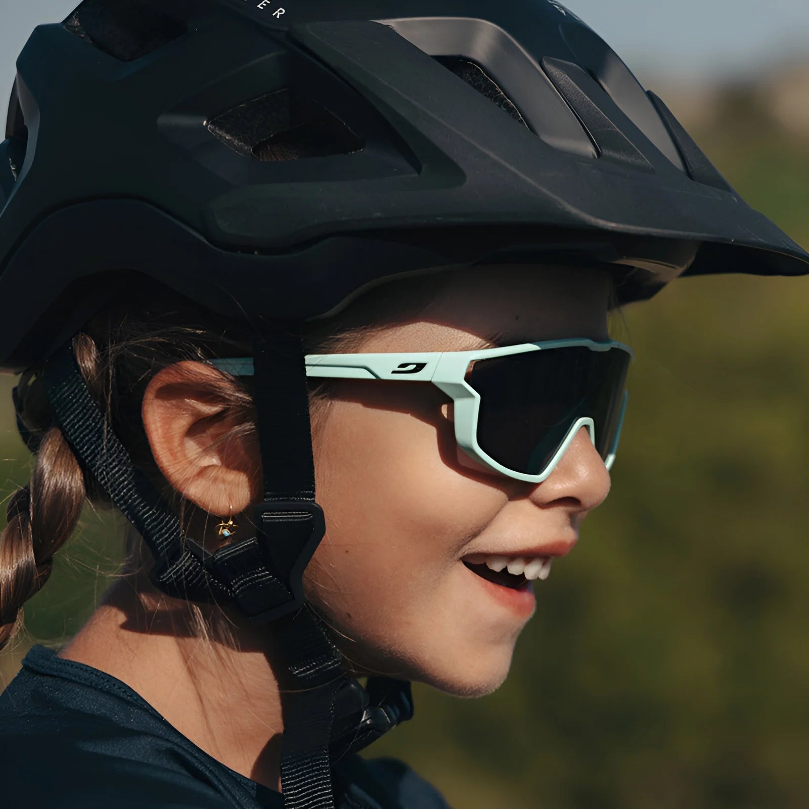 A child, wearing a cycling helmet and Julbo's Fury Mini sunglasses with comfortable side venting, smiles broadly while enjoying the outdoors.