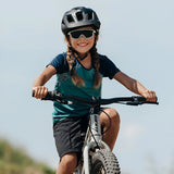A braided child grins joyfully while cycling outdoors, relishing the panoramic view through their Julbo Fury Mini sunglasses by Julbo, which feature side venting for ultimate comfort.