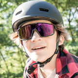 A person wearing a red plaid shirt and the Julbo Fury S Sunglasses with a helmet enjoys the outdoors. The sunglasses offer a wide field of vision, making every adventure more immersive and vivid.