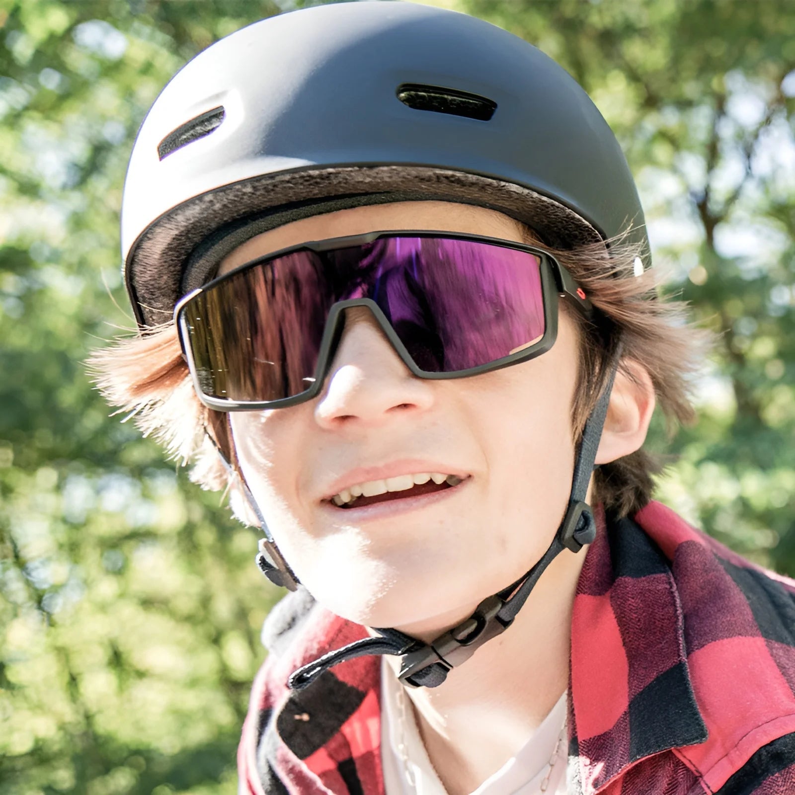 A person wearing a red plaid shirt and the Julbo Fury S Sunglasses with a helmet enjoys the outdoors. The sunglasses offer a wide field of vision, making every adventure more immersive and vivid.