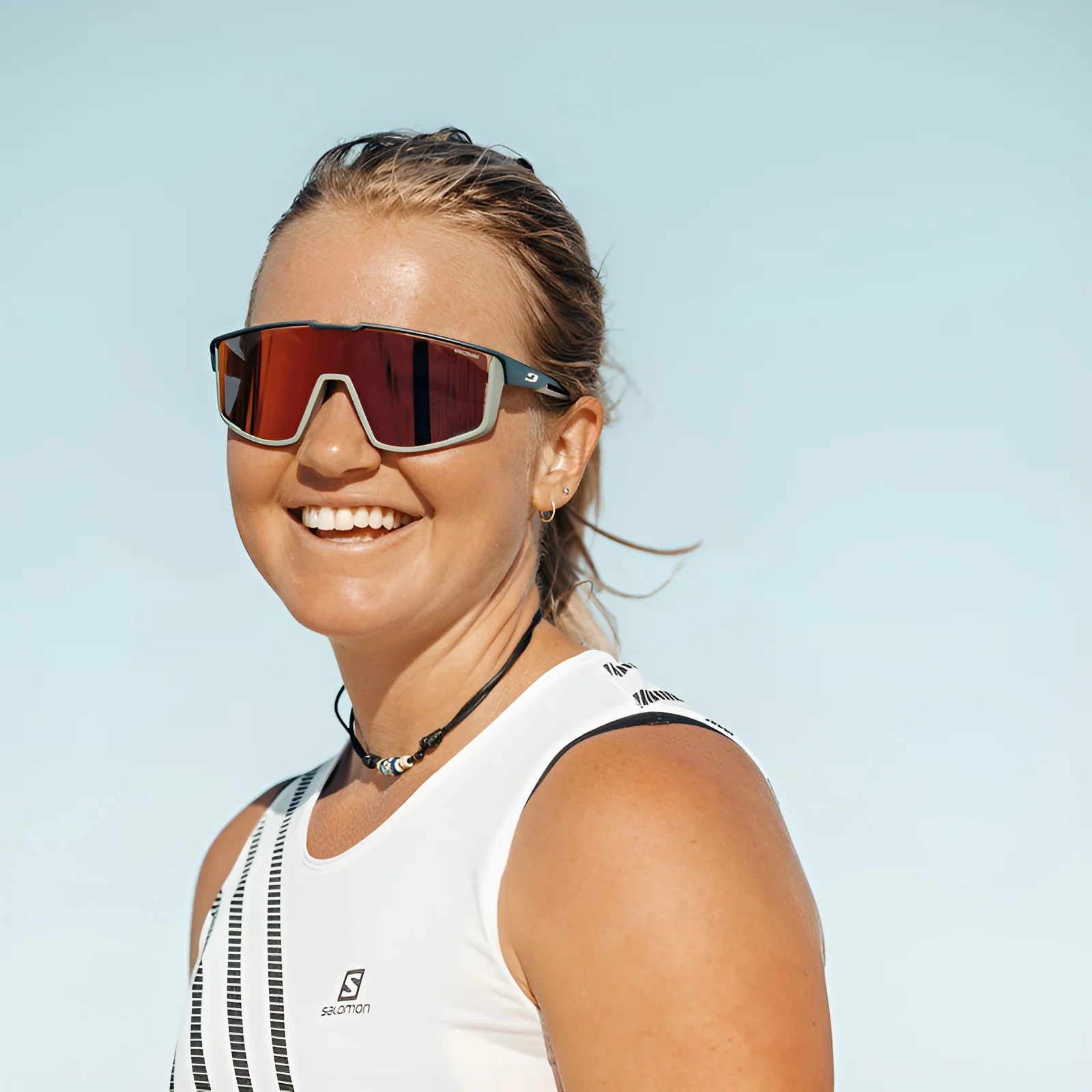 A woman smiles in Julbo Fury sunglasses with REACTIV photochromic lenses and a white tank top under a clear blue sky.