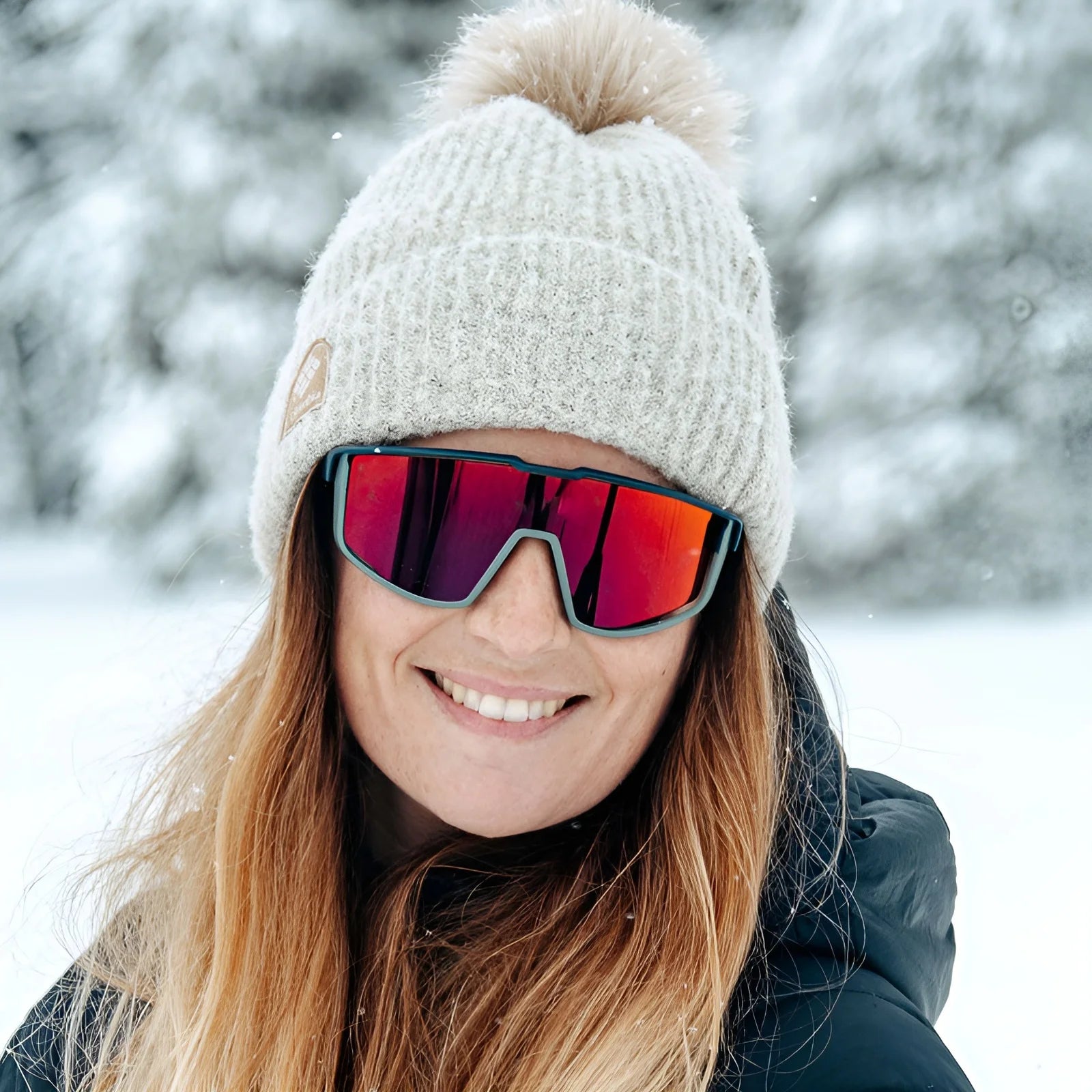 A woman in a snowy landscape wears Julbo Fury sunglasses with REACTIV photochromic lenses, coupled with a beige knit hat and the Air Link Temple System for comfort and style.