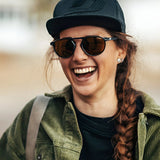 A woman with braided hair and a cap smiles confidently, showcasing her urban look. She's wearing a green jacket and the Julbo Meta Sunglasses, size 55, adding to her style.