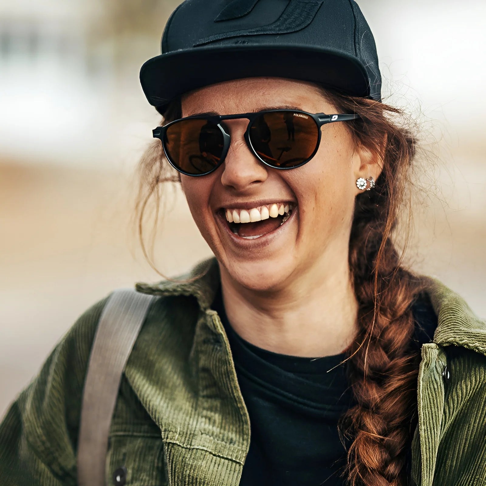 A woman with braided hair and a cap smiles confidently, showcasing her urban look. She's wearing a green jacket and the Julbo Meta Sunglasses, size 55, adding to her style.