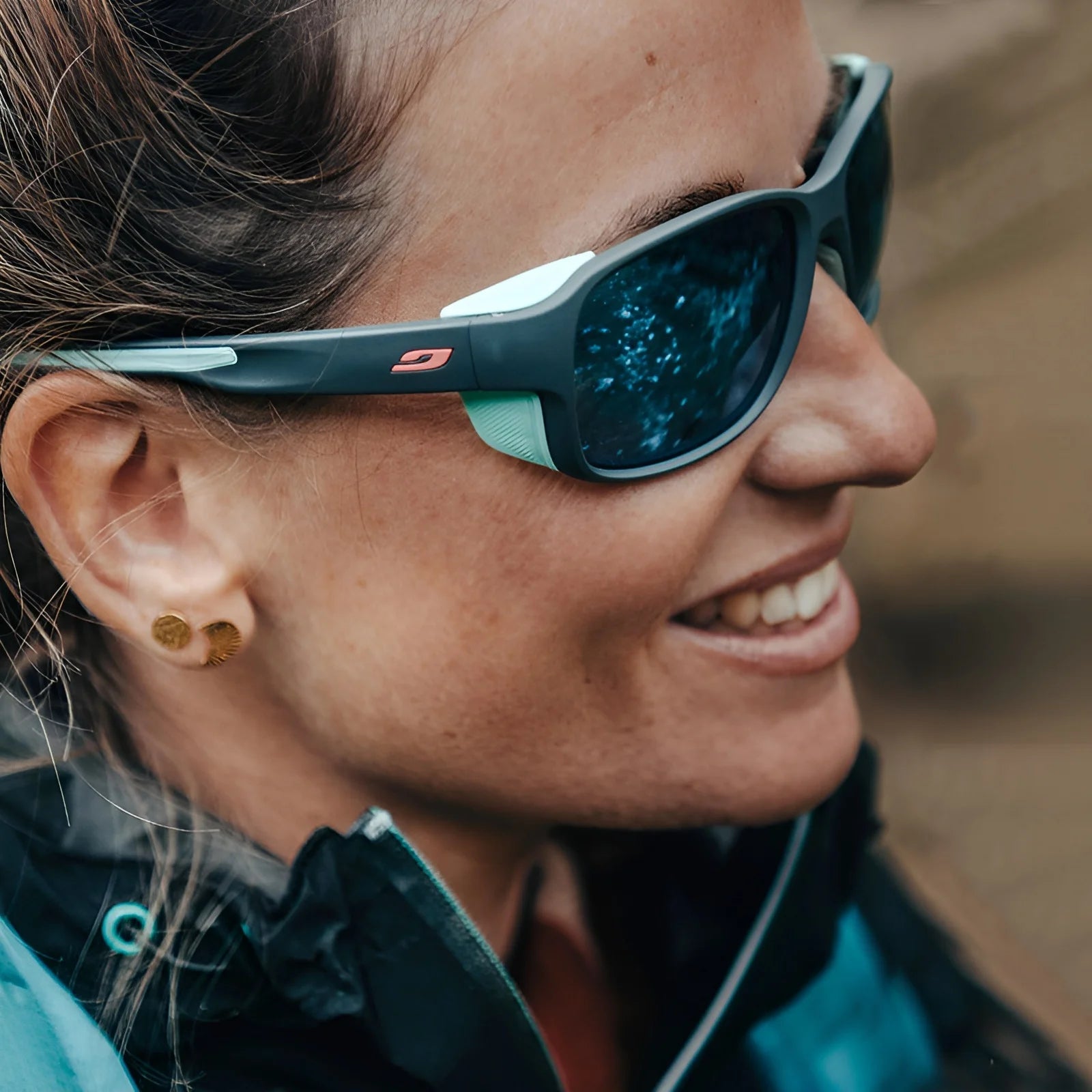A smiling woman, with hair tied back and gold earrings, wears Julbo Monterosa 2 Sunglasses Size 54 in a protective design for mountain comfort along with a jacket.