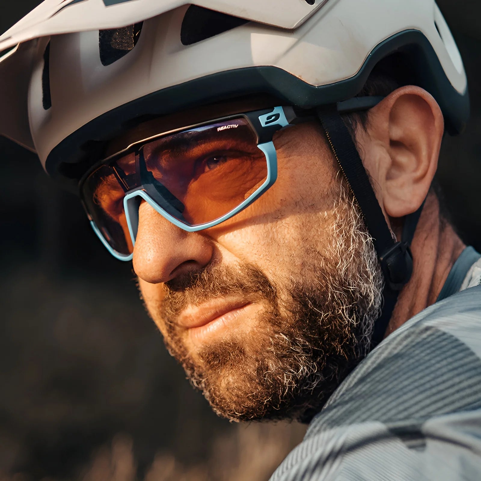 A man in a helmet and Julbo Rush Sunglasses, featuring orange-tinted, photochromic lenses, gazes thoughtfully into the distance.