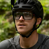 A person wearing a black bike helmet and Julbo Rush Sunglasses with adjustable temples, looking up against a green blurred background.