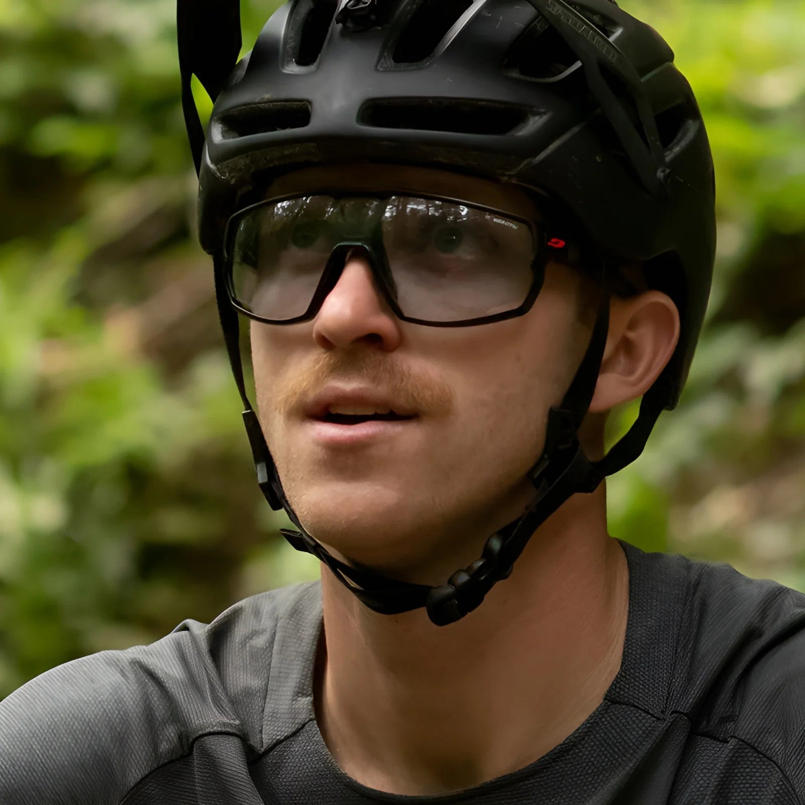 A person wearing a black bike helmet and Julbo Rush Sunglasses with adjustable temples, looking up against a green blurred background.