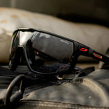 The Julbo Rush Sunglasses, featuring black and red frames with adjustable temples, rest on a dark surface, reflecting outdoor scenery.