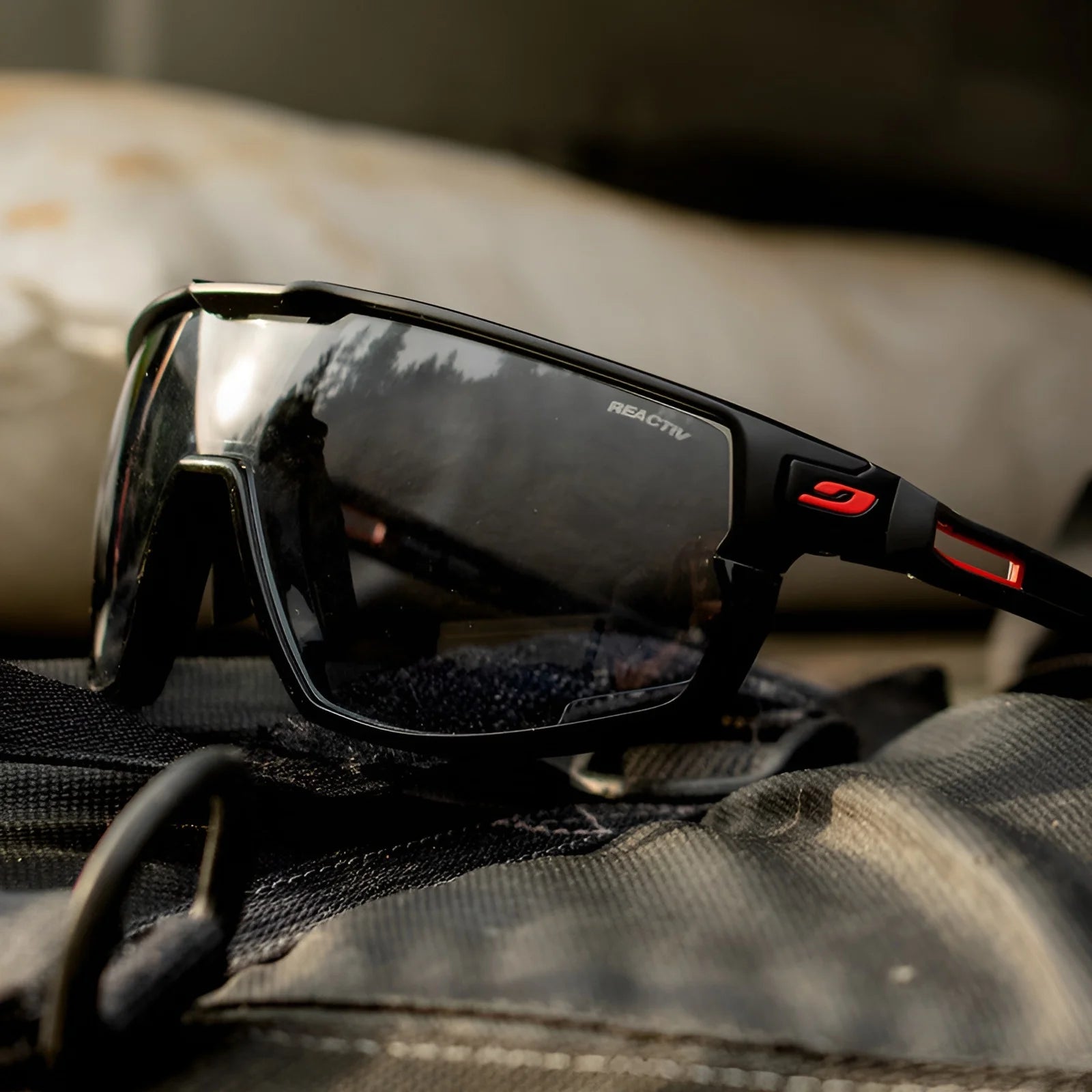 The Julbo Rush Sunglasses, featuring black and red frames with adjustable temples, rest on a dark surface, reflecting outdoor scenery.
