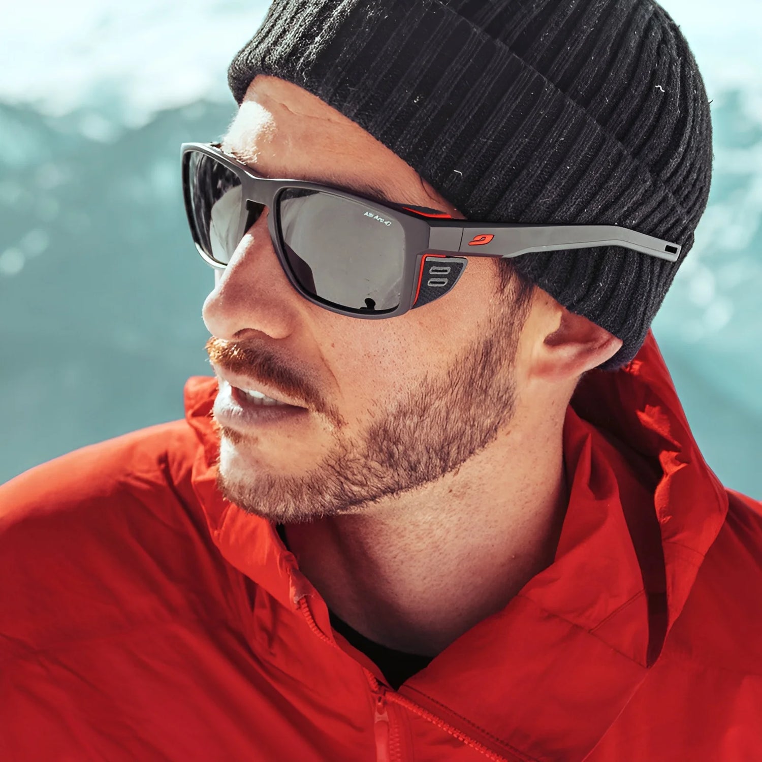 Man in a red jacket, black beanie, and Julbo Shield Sunglasses (Size 59) facing left against a snowy background.