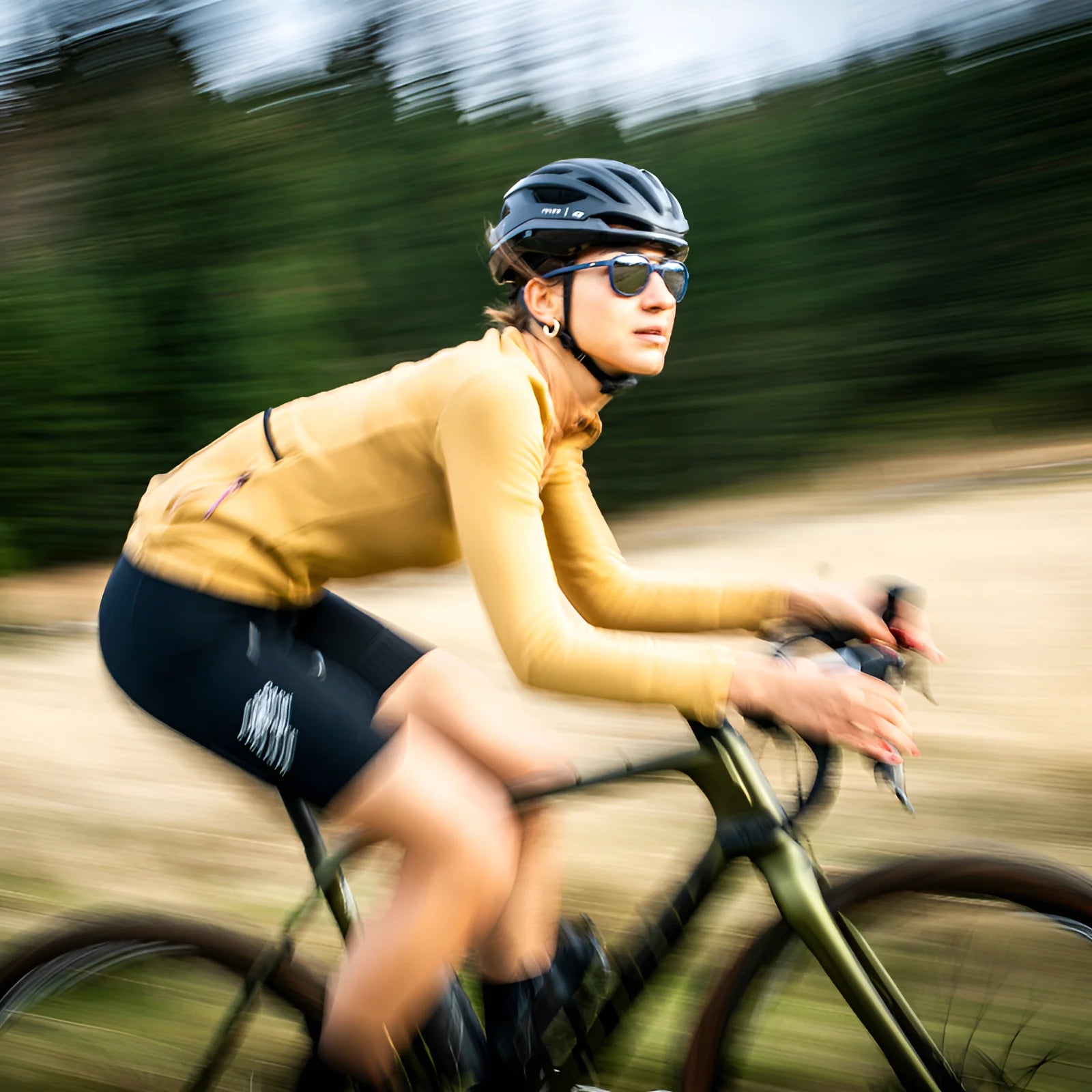 A person, wearing a vibrant yellow jacket and cycling gear with Julbo Slack Sunglasses (Size 52), rides swiftly through lush greenery, the trees blurring as they pass.