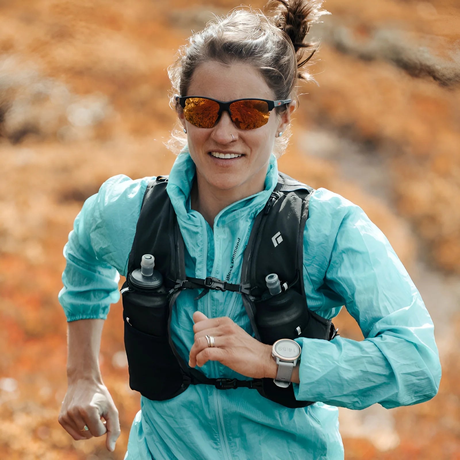 A person jogs through an autumn landscape in a blue jacket and hydration vest, wearing sleek Julbo Split Sunglasses | Size 63 with REACTIV photochromic lenses adapting effortlessly to changing light.