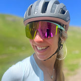 A person smiles wearing Julbo The Bright Series Sunglasses and a cycling helmet, with a green landscape in the background.