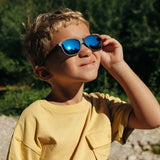 A young boy wearing Julbo Turn 2 Sunglasses (Size 45) and a yellow shirt squints in the sun, surrounded by a lush, green outdoor setting.