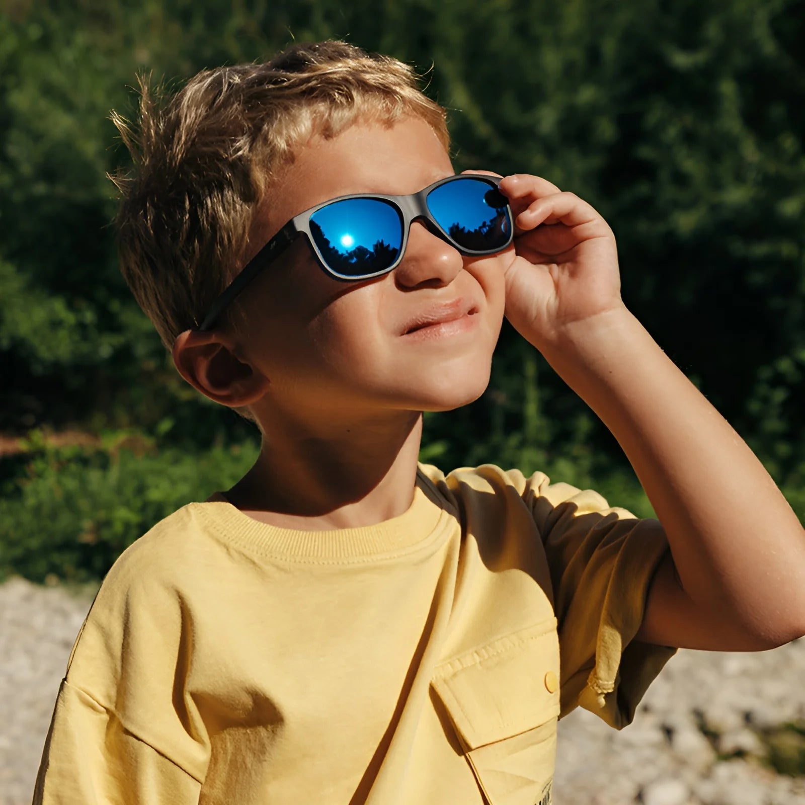 A young boy wearing Julbo Turn 2 Sunglasses (Size 45) and a yellow shirt squints in the sun, surrounded by a lush, green outdoor setting.