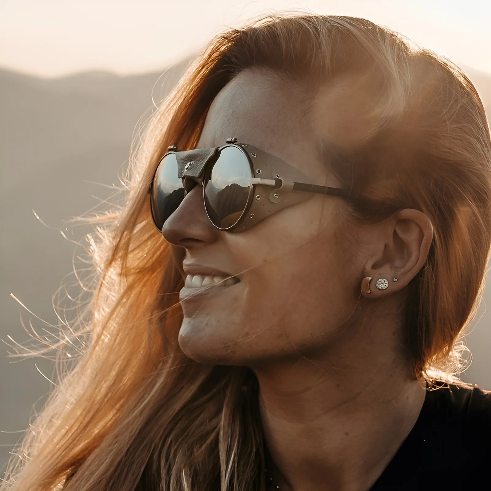 A woman with long hair smiles outdoors, wearing Julbo Vermont Classic Sunglasses (Size 51), which reflect the majestic mountain backdrop.