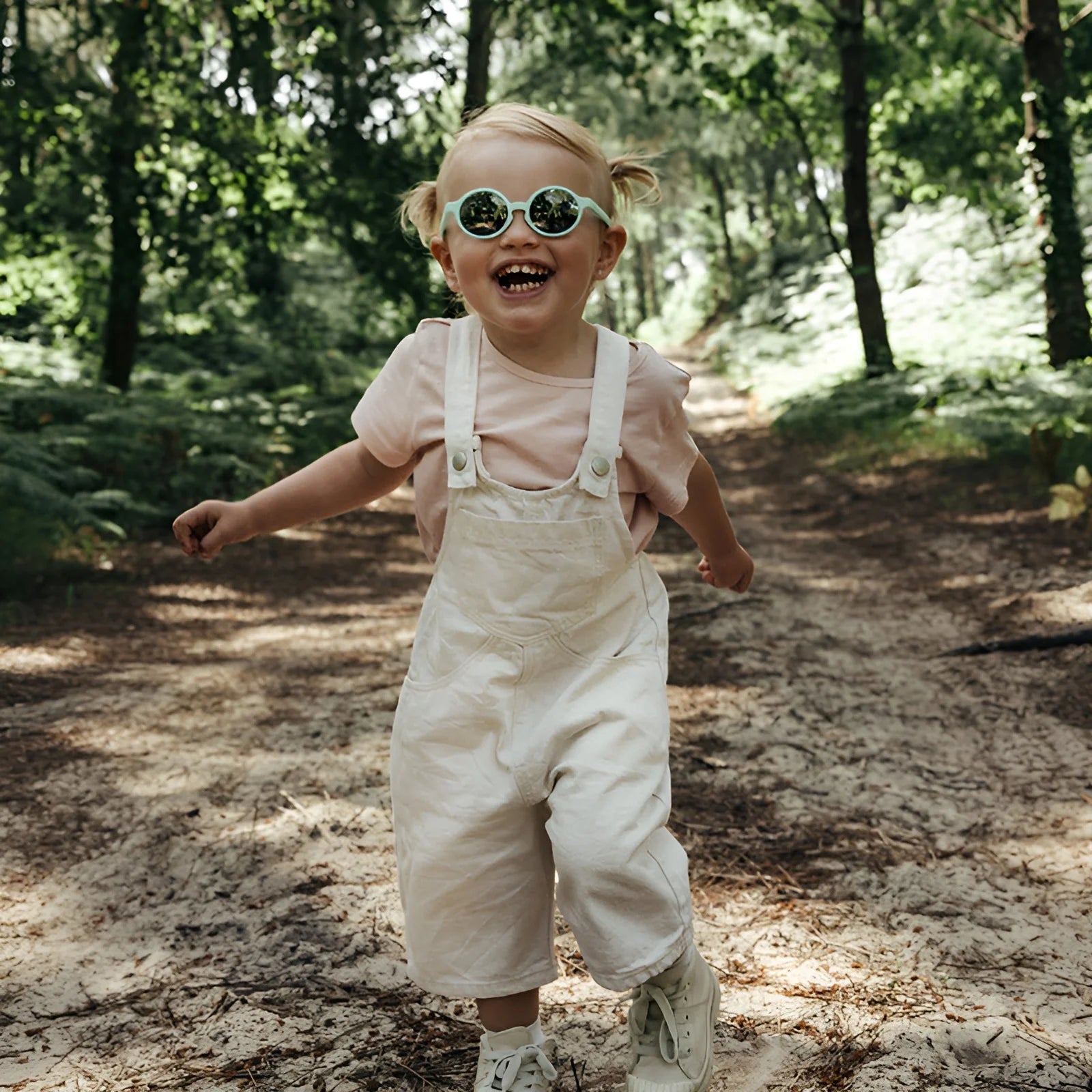A young child with Julbo Walk Sunglasses and pigtails joyfully runs down a sunlit forest path, wearing a pink shirt and white overalls.